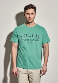 Jasnozielona bawełniana koszulka z krótkim rękawem, z granatowym napisem "LE SOLEIL - TOUTE L'ANNÉE - Port Fleur" na przodzie.
