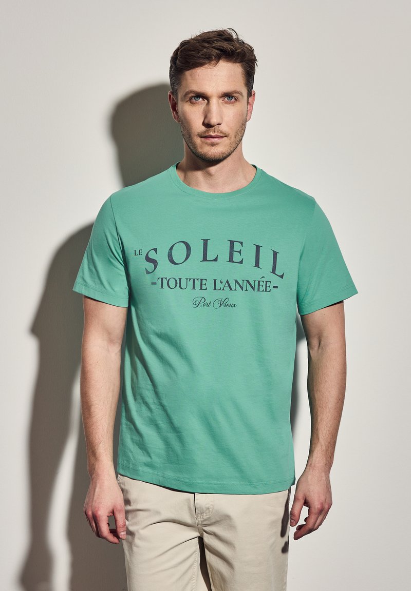 Jasnozielona bawełniana koszulka z krótkim rękawem, z granatowym napisem "LE SOLEIL - TOUTE L'ANNÉE - Port Fleur" na przodzie.