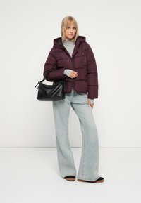 Chaqueta acolchada de color púrpura oscuro con capucha, combinada con unos vaqueros de campana de color azul claro. El modelo sostiene un bolso negro texturizado con un asa fruncida.