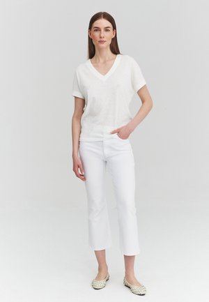 Donna con capelli castani lisci che indossa una maglia bianca con scollo a V e maniche corte, pantaloni bianchi corti e ballerine beige con motivo, in piedi davanti a uno sfondo neutro.