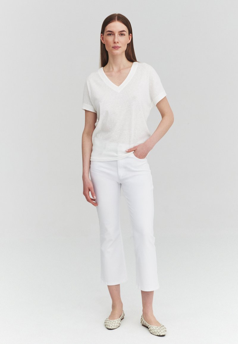 Femme aux cheveux bruns raides portant un haut blanc à col en V et manches courtes, un pantalon blanc court et des ballerines beige à motifs, debout devant un fond uni.