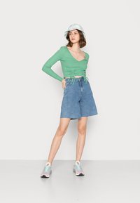 Blusa longa de manga com nervuras verdes e detalhe de laço na cintura, combinada com calções de ganga de cintura alta e ténis pastéis, complementada por um leve chapéu de balde.