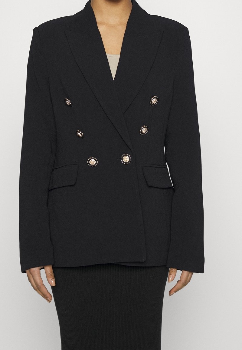 Personne portant un blazer noir croisé avec des boutons dorés et des poches à rabat par-dessus un haut beige et une jupe noire.