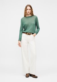 Žena s dlhými vlnitými vlasmi, oblečená v zelenej mikine, bielych širokých nohaviciach s hnedým opaskom a hnedými členkovými topánkami, stojaca s jednou rukou v vrecku.