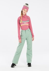Tenue de ski comprenant un haut ajusté à manches longues rose avec des accents de motifs et un pantalon de neige vert clair; porté avec un bonnet beige et vert à pompon.