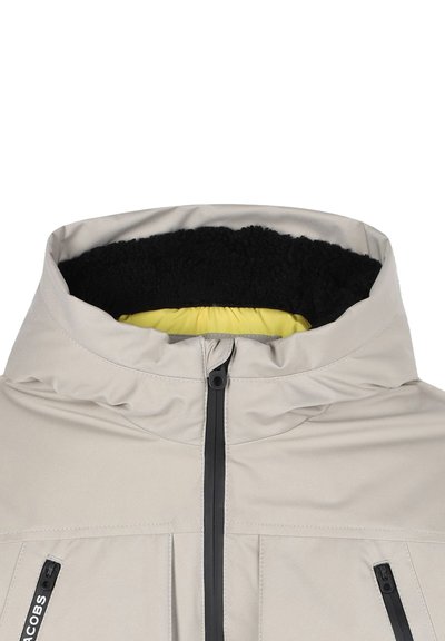 Veste beige clair avec un col montant, doublure en polaire noire et intérieur jaune. Présente une fermeture éclair à l'avant et deux poches latérales.