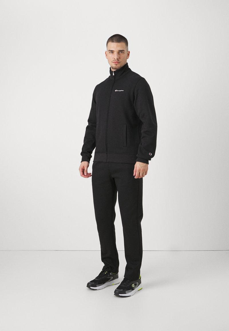 Chándal deportivo negro de tejido suave, con chaqueta con cremallera y bolsillos laterales, y pantalones a juego, ambos con detalle de logo.