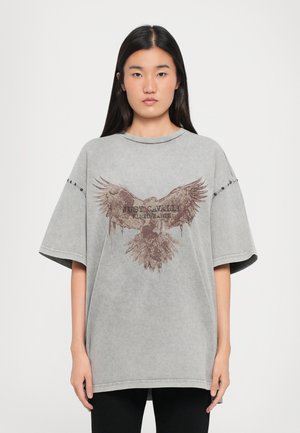 T-shirt oversize gris en tissu doux, arborant un grand graphisme d'aigle et le texte "JUST CAVALLI KID EARTH" en marron. Manches courtes et col rond.
