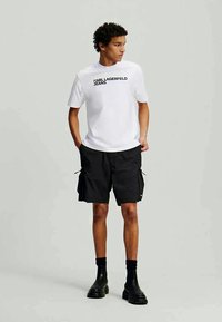 Jovem com cabelo encaracolado a usar uma t-shirt branca da Karl Lagerfeld Jeans, calções cargo pretos, meias pretas e botas grossas pretas.