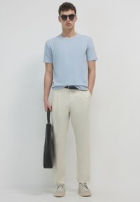 T-shirt azzurro chiaro a maniche corte, pantaloni crema, scarpe beige e borsa a tracolla nera. Gli occhiali da sole completano l'outfit, mantenendo un aspetto informale.