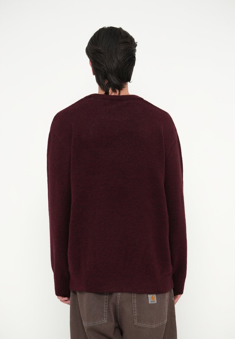 Bordeaux gebreide trui met een relaxte pasvorm, geribbelde halslijn en lange mouwen, gecombineerd met bruine broek met een merklabel op de achterkant.