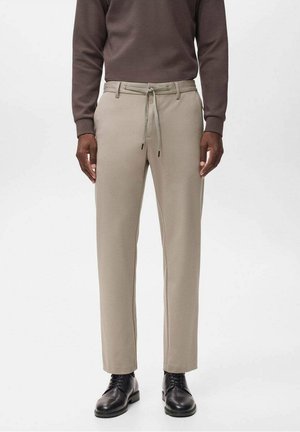 Man die beige broek met trekkoord draagt, zwarte leren veterschoenen en een bruin overhemd met lange mouwen, staand tegen een effen achtergrond.
