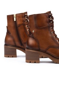 Botas de cuero marrón hasta el tobillo con cremallera lateral, puntera redonda y un tacón bloque texturizado. Presentan ojetes metálicos y un detalle de costura a lo largo de la suela.