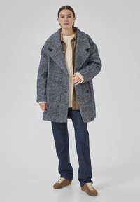 Žena oblečená v modro-bielej herringbone kabáte, pod ktorým má béžový sveter a hnedú košeľu, v tmavých džínsoch a pieskových teniskách, stojí na šedom pozadí.