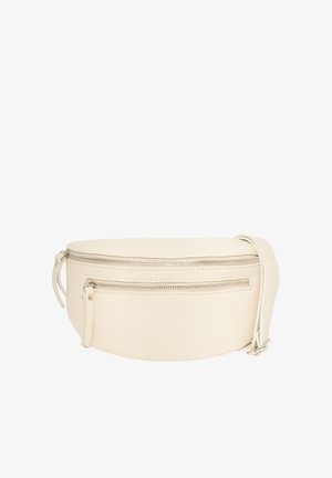 Frau trägt ein schwarzes Langarmshirt und dunkle Jeans mit einer beigen Umhängetasche, die auf ihrer Brust liegt, und hat die Hände in den Taschen.