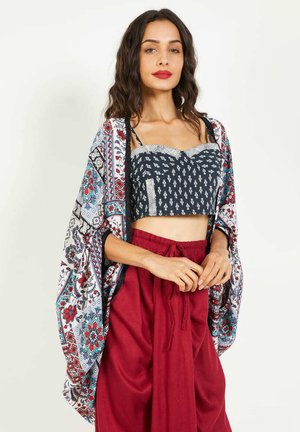 Mujer con cabello largo y oscuro, usando lápiz labial rojo, una blusa recortada azul con estampado, un chal floral colorido y pantalones rojos de tiro alto y holgados, de pie.