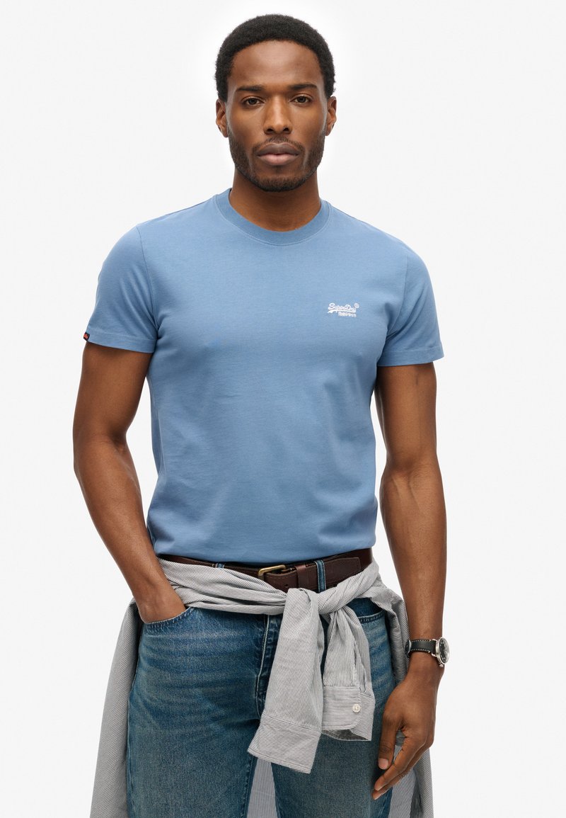 Man in een blauw T-shirt en jeans met een gestreepte overhemd om zijn taille gebonden, handen in de zakken, met een horloge om zijn pols, staande tegen een witte achtergrond.