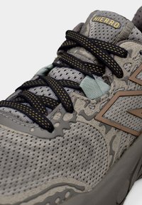 Αθλητικό παπούτσι ανοιχτού γκρι με επάνω μέρος από δίχτυ, καφέ λεπτομέρειες και μαύρα κορδόνια με κίτρινες βούλες. Το σχέδιο έχει υφή με επικαλυπτόμενα στρώματα.