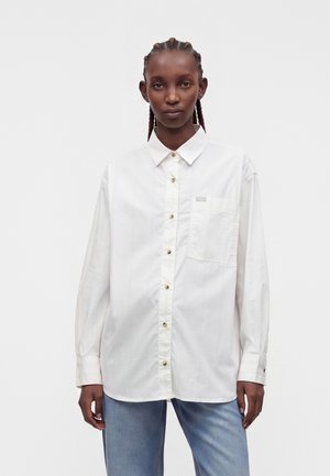Junge Person mit geflochtenem Haar trägt ein übergroßes weißes Hemd mit Knöpfen und hellblaue Jeans vor einem schlichten weißen Hintergrund.