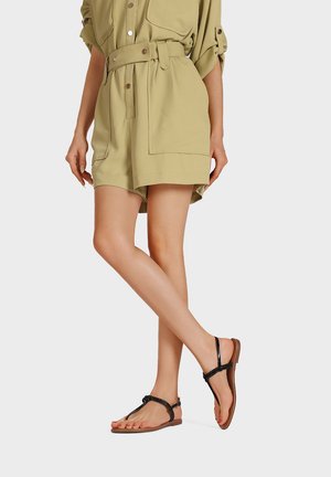 Donna che indossa pantaloncini beige con cintura e sandali neri piatti con cinturino a T, in piedi con una gamba incrociata sull'altra contro uno sfondo uniforme.