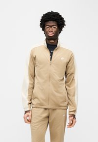 Μπεζ ζακέτα με φερμουάρ και λευκά μανίκια, κατασκευασμένη από λείο ύφασμα. Διαθέτει πλαϊνές τσέπες και μικρό λευκό λογότυπο Nike στο στήθος.