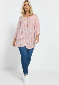 Vaaleanpunainen paisley-kuosiset tunika, jossa on kolme neljänneksen hihaa, pyöreä pääntie ja nyöritys, yhdistettynä sinisiin skinny-farkkuihin ja valkoisiin lenkkareihin.