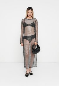 Vestido longo de malha preta, semi-transparente sobre um biquíni preto. A modelo segura uma pequena bolsa preta; os sapatos têm biqueiras em ponta e laços.