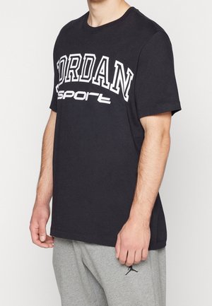 Man draagt een zwart Jordan sport T-shirt en lichtgrijze joggingbroek met klein Jordan-logo op het bovenbeen, staand tegen een effen achtergrond.