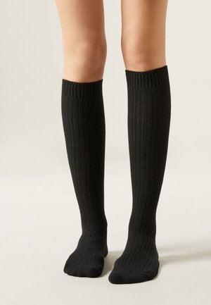 Piernas con calcetas negras de canalé hasta la rodilla, de pie sobre un fondo claro y neutro, con los pies relajados.