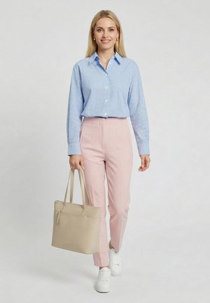 Kobieta idąca, ubrana w niebieską koszulę w kratę, różowe spodnie, białe trampki i niosąca beżową torbę na jasnym jednolitym tle.