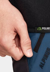 Czarna dzianina na górze z zieloną metką "POLARIS"; poniżej niebieskie spodnie z ciemnym akcentem i wytłoczonym tekstem "ALPINUS". Ręka chwyta bok.