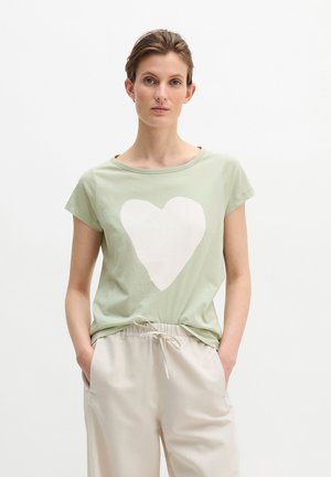 Kobieta w jasnooliwkowej koszulce z dużym białym sercem na przodzie, włożonej do beżowych spodni z troczkiem, z rękami w kieszeniach.