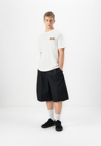 Ung mann iført hvit Rip Curl Australia t-skjorte, løse svarte shorts til knærne, hvite sokker og svarte joggesko, stående mot en hvit bakgrunn.