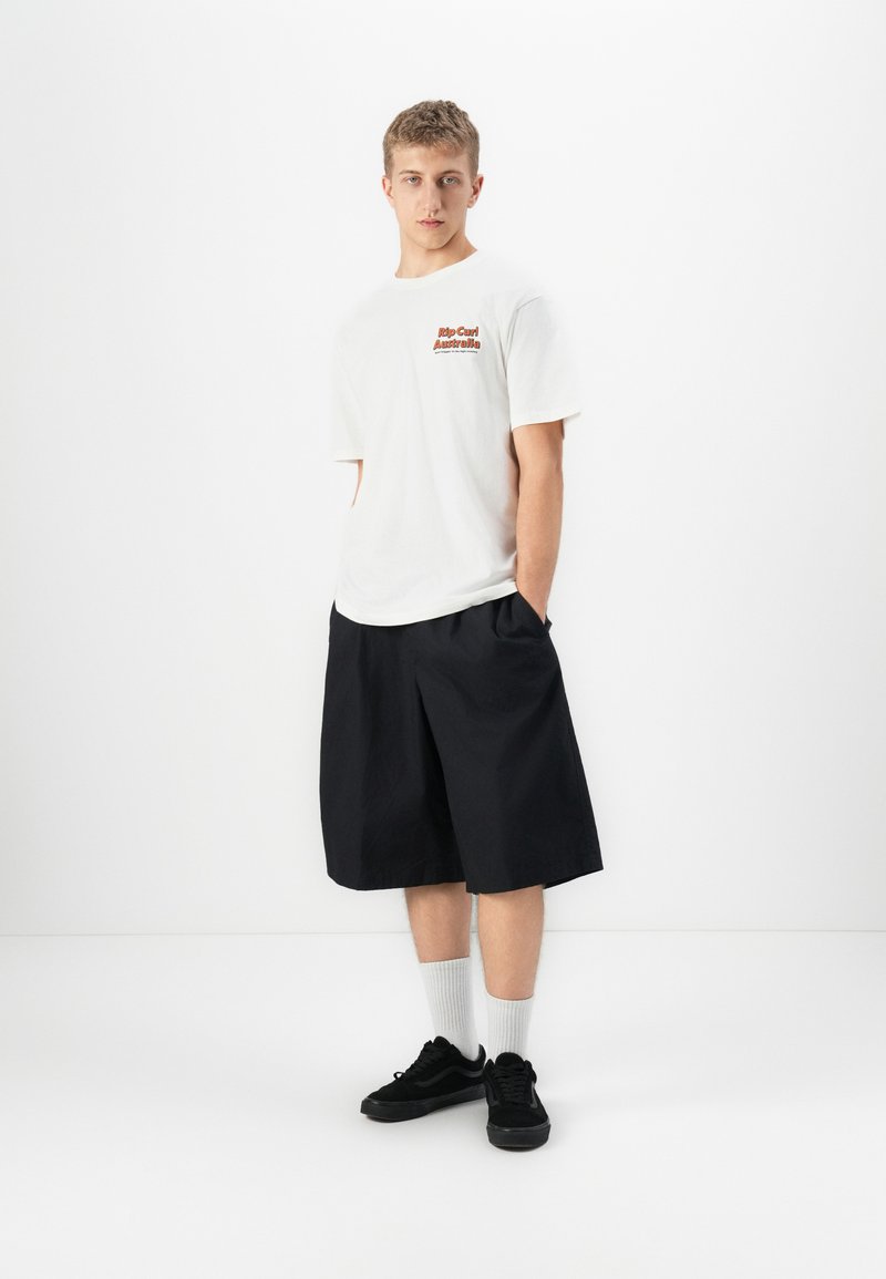 Ung mann iført hvit Rip Curl Australia t-skjorte, løse svarte shorts til knærne, hvite sokker og svarte joggesko, stående mot en hvit bakgrunn.