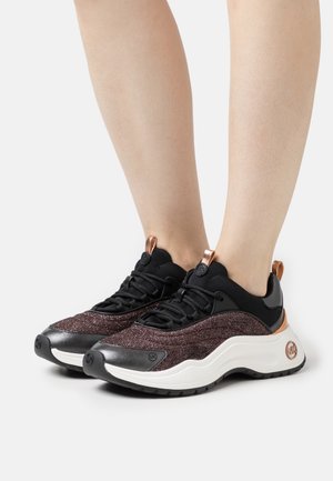 Pieds portant des baskets noires et violet foncé scintillantes avec semelles blanches et accents en or rose, posés sur un fond blanc uni.