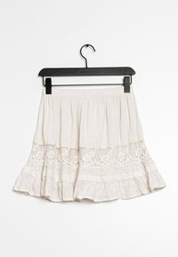 Jupe à volants beige clair en tissu léger, ornée de broderies florales et de dentelle au niveau de l'ourlet. Suspendue à un crochet noir.