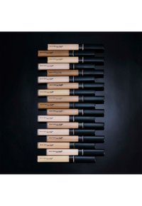 Une rangée de tubes de correcteur Maybelline dans des teintes variées, avec une finition mate, des bouchons noirs et des corps transparents affichant la teinte du produit.