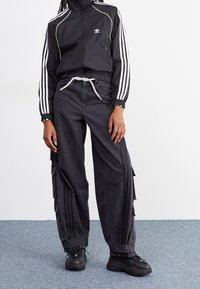 Svart träningsjacka med vita ränder och gröna accenter, kombinerad med lösa svarta cargobyxor med blixtlås och sidofickor. Svarta sneakers.