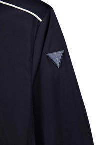 Marinejacke mit weißem Paspel, versehen mit einem dreieckigen blauen Logopatch auf dem linken Ärmel. Glatte Stoffoberfläche, modernes Design.