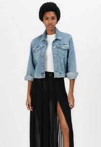 Giacca di jeans in un blu chiaro, dal design cropped, con chiusura a bottoni, tasche sul petto e maniche arrotolate. Abbinata a una gonna nera fluida.