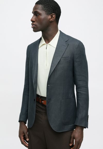 Άνδρας που φοράει σκούρο μπλε σακάκι, κρεμ πουκάμισο με φερμουάρ, καφέ παντελόνι και καφέ ζώνη, στέκεται μπροστά σε απλό ανοιχτό φόντο κοιτάζοντας πλάγια.