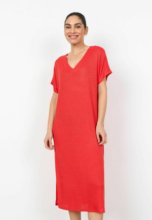 Ženska se smeji, nosi široko, do kolen dolgo rdečo obleko s kratkimi rokavi in V-izrezom, stoji pred belim ozadjem.