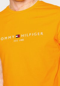 Camiseta de algodón naranja con un logo blanco "TOMMY HILFIGER" y "EST. 1985" en una fuente limpia, con un pequeño emblema de color en el centro.