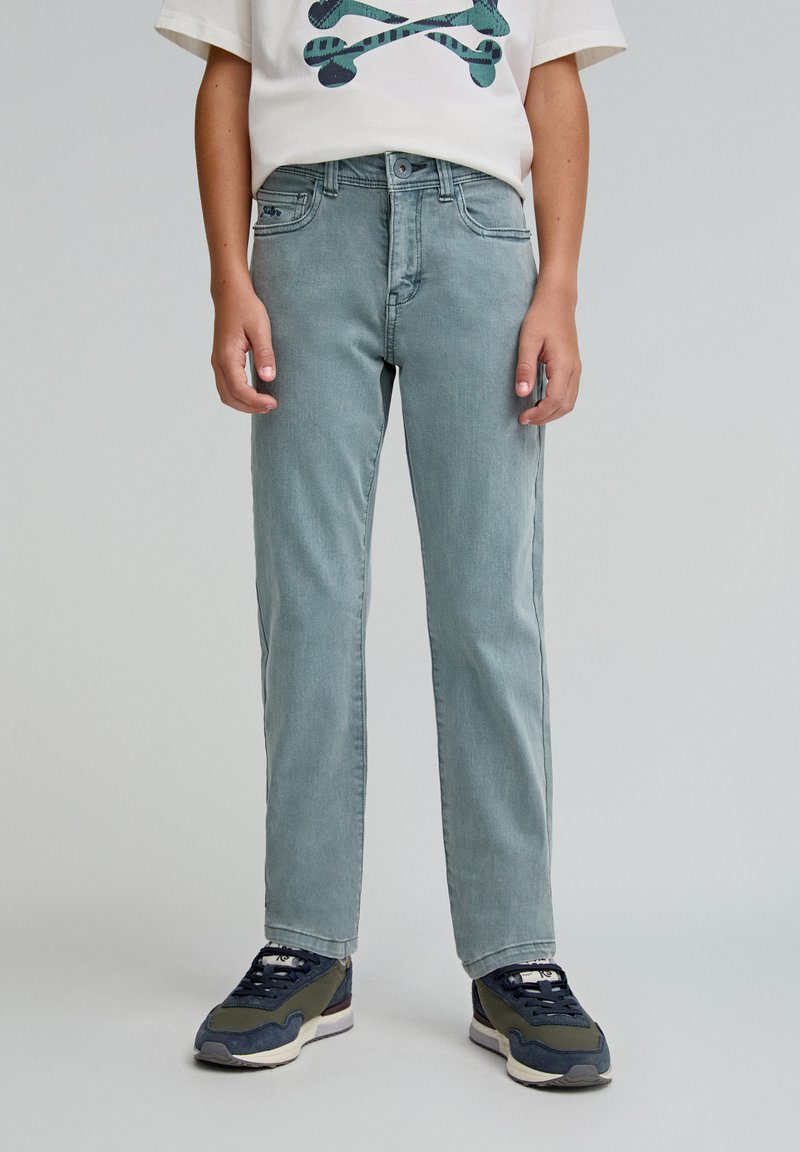 Jeans de denim azul claro con corte recto, diseño de cinco bolsillos y un sutil efecto de desgarro. Combinados con zapatillas oscuras y una camiseta gráfica blanca.