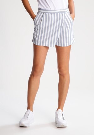 Femme portant un short à rayures verticales blanches et grises, des baskets blanches, et un haut blanc avec les mains dans les poches.