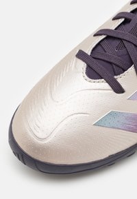 Chaussure de soccer beige clair avec une tige texturée et brillante, accents violets, détails perforés et semelle en caoutchouc. Présente des motifs en relief.