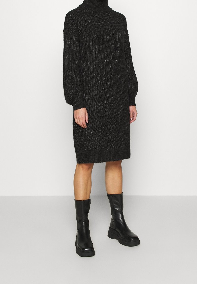 Personne portant une robe en tricot noir à col roulé avec des manches ballon et des bottes en cuir noires midi-cheville, se tenant sur un sol gris uni.