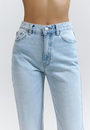 Personne portant un jean en denim bleu clair taille haute avec bouton devant et poches, montré de la taille jusqu'au milieu de la cuisse sur un fond uni.