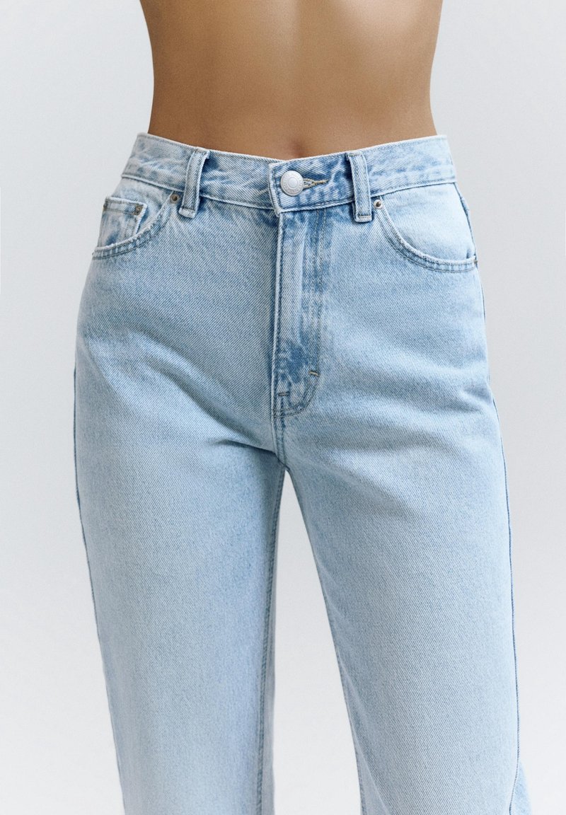 Personne portant un jean en denim bleu clair taille haute avec bouton devant et poches, montré de la taille jusqu'au milieu de la cuisse sur un fond uni.
