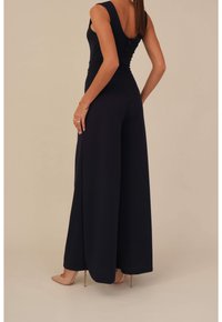 Mujer con un mono negro sin mangas y pierna ancha, usando tacones nude, de pie con las manos en los costados sobre un fondo beige.